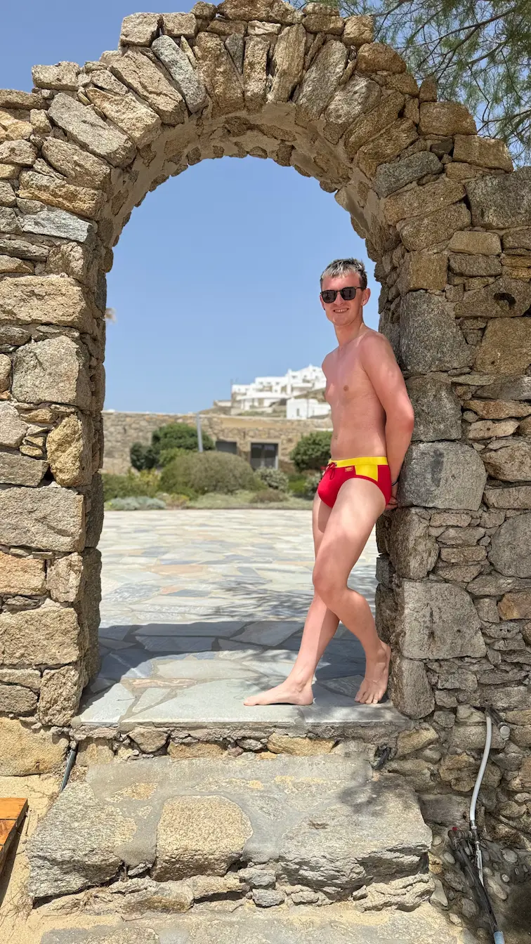 Alex in red speedos on the Elia Beach at the Beach Club under a stone arch.